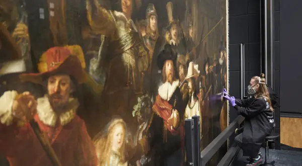 Restauratie Nachtwacht: een vrouw met mondkapje en handschoenen werkt aan het beroemde schilderij van Rembrandt.