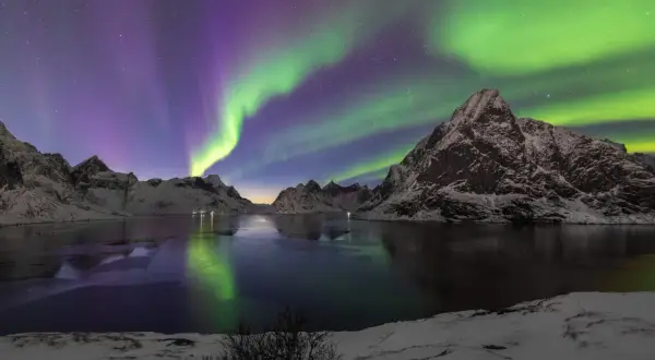 Spectaculaire foto's van het noorderlicht boven besneeuwde bergen en een bevroren meer. De groene en paarse gloed reflecteert in het water.