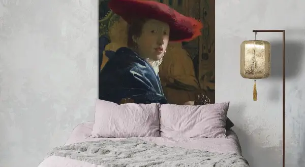 Een meisje met de rode hoed, geschilderd door Vermeer, kijkt mysterieus vanaf de muur boven een zachtroze bed.