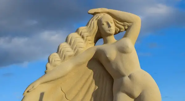 Zandstenen beeld van de Griekse godin Aphrodite, met golvend haar en een hand op haar hoofd, tegen een blauwe lucht met wolken.