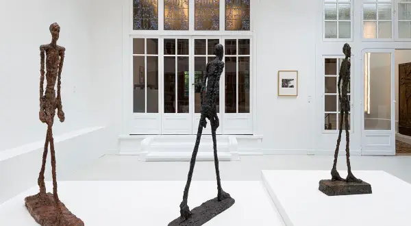 Drie slanke, bronzen beelden van l'homme qui marche staan in een witte galerie. De figuren lijken te bewegen, met hun lange benen en dunne lichamen.