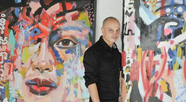 Peter Klashorst staat naast zijn kleurrijke, abstracte schilderijen. Een portret met een oog en rode lippen, en een ander werk met veel lijnen en verfspatten.