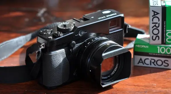 Zwarte Fujifilm X-E1 camera met lenskap en filmrolletjes Acros 100 op een houten tafel.