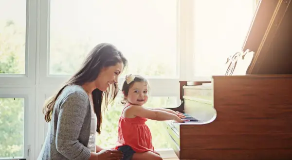 Een moeder helpt haar dochtertje met het spelen op de piano. Het kleine meisje zit op de pianokruk en lacht vrolijk terwijl ze de toetsen aanraakt.