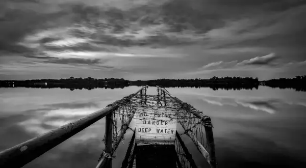 Zwart-wit fotograaf legt een verlaten pier vast met een waarschuwingsbord "DANGER DEEP WATER". De weerspiegeling in het water is sereen.