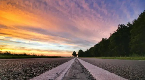 Een lege weg met witte lijnen, gefotografeerd vanuit ooghoogte perspectief. De lucht kleurt oranje en paars bij zonsondergang, met bomen aan de rechterkant.