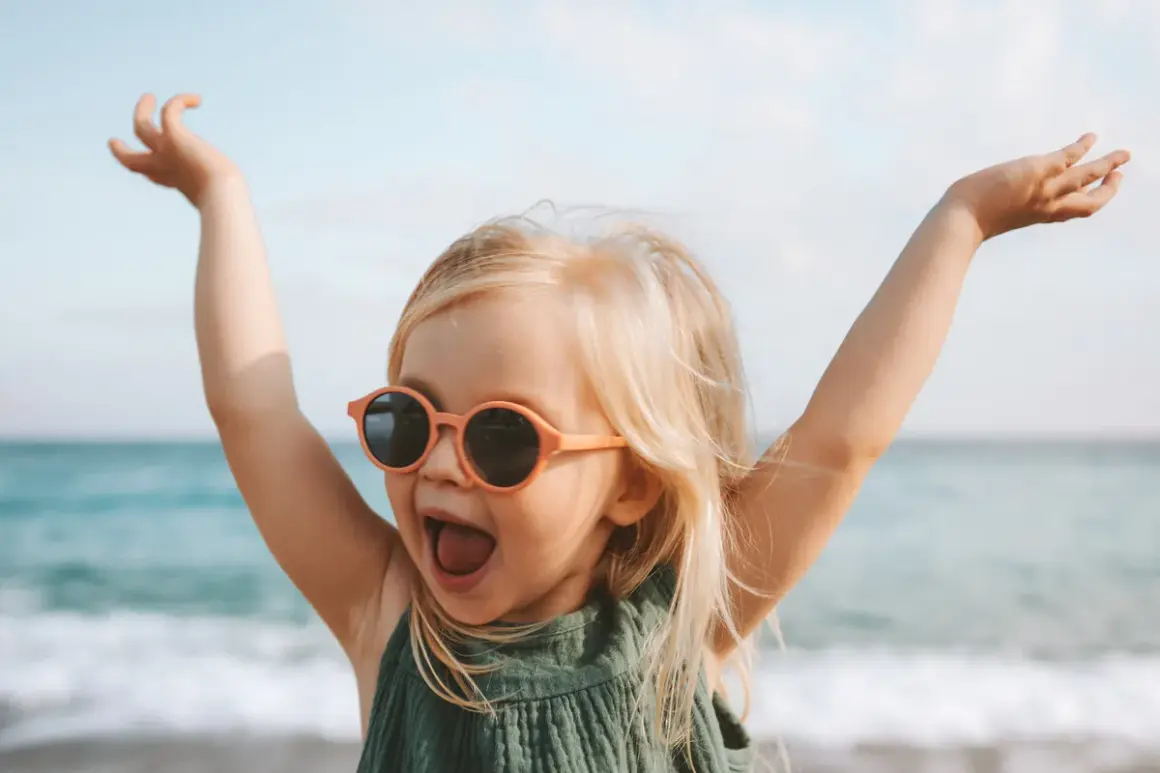 Vrolijk meisje met zonnebril op het strand, armen in de lucht. Ze lijkt wel een kleine godin, met namen als Freya of Aphrodite.