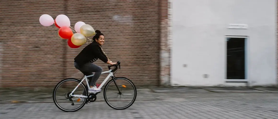 Vrolijke vrouw op een fiets met ballonnen, bewegende mensen in een stedelijke omgeving.