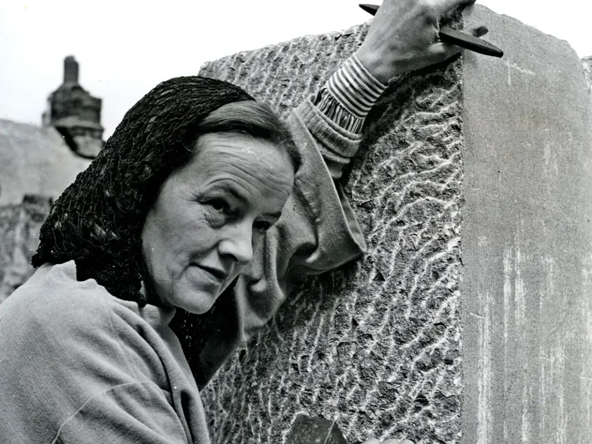 Barbara Hepworth, met een hoofddoek, bewerkt een steen met een beitel. Haar handen tonen de concentratie van een kunstenaar.