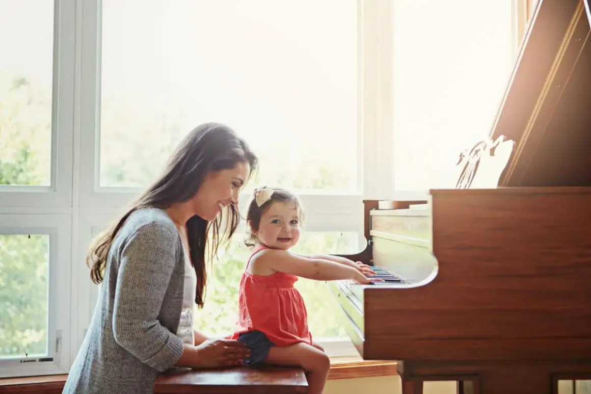 Een moeder helpt haar dochtertje met het spelen op de piano. Het kleine meisje zit op de pianokruk en lacht vrolijk terwijl ze de toetsen aanraakt.