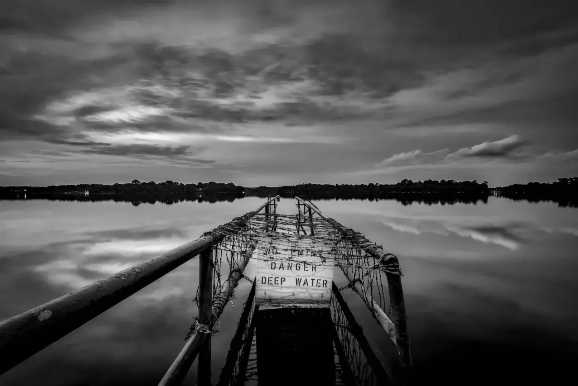 Zwart-wit fotograaf legt een verlaten pier vast met een waarschuwingsbord "DANGER DEEP WATER". De weerspiegeling in het water is sereen.