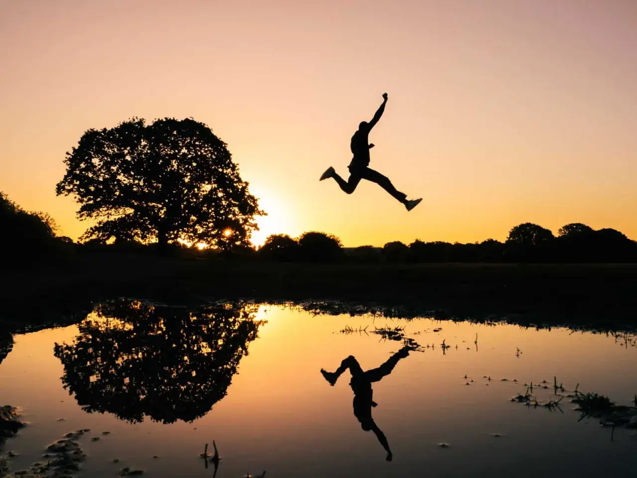 Silhouet van een persoon die springt bij zonsondergang, met een reflectie in het water. Een perfecte burst foto Samsung.