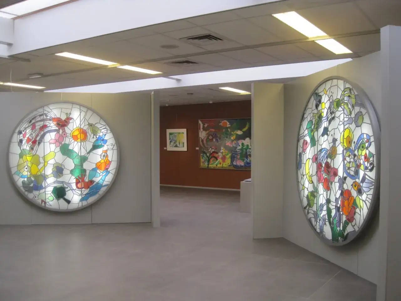 Twee ronde glas-in-loodwerken, mogelijk van Hans Truijen, sieren de muren van een galerie. Op de achtergrond hangen kleurrijke schilderijen.