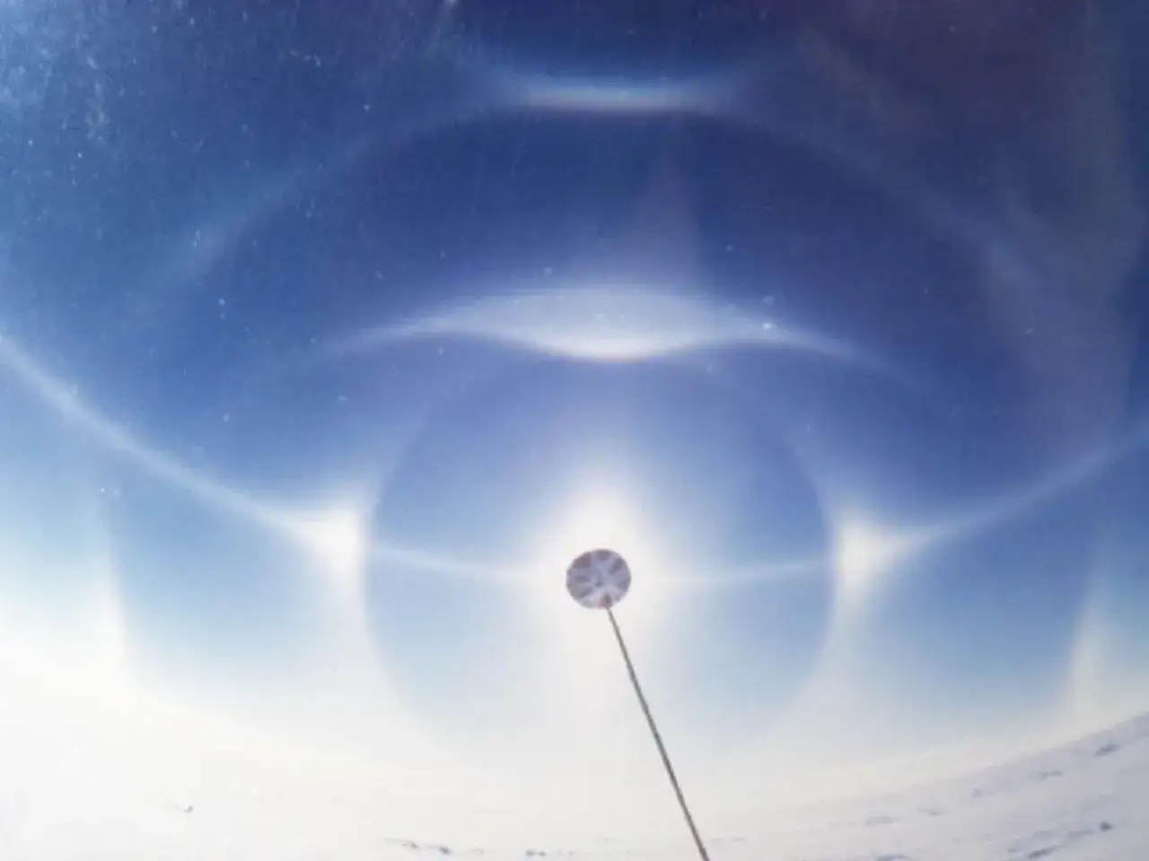 Een spectaculaire halo effect zon met heldere cirkels en bijzonnen boven een besneeuwd landschap. Een instrument steekt omhoog.