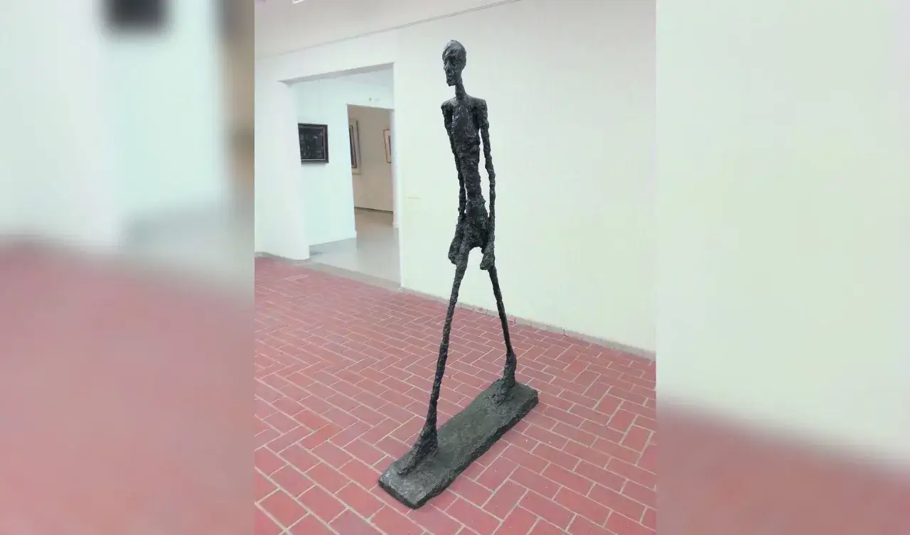 L'homme qui marche, een slanke, bronzen figuur, staat op een tentoonstellingsvloer met rode bakstenen.