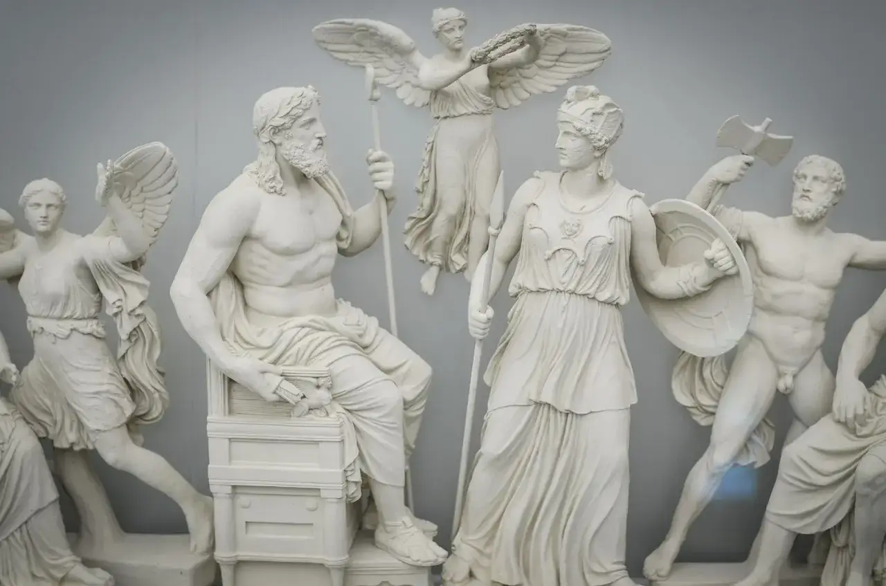 Wit marmeren beeldengroep met godinnen namen, waaronder een zittende man, een krijgsgodin met schild en speer, en een gevleugelde figuur met lauwerkrans.