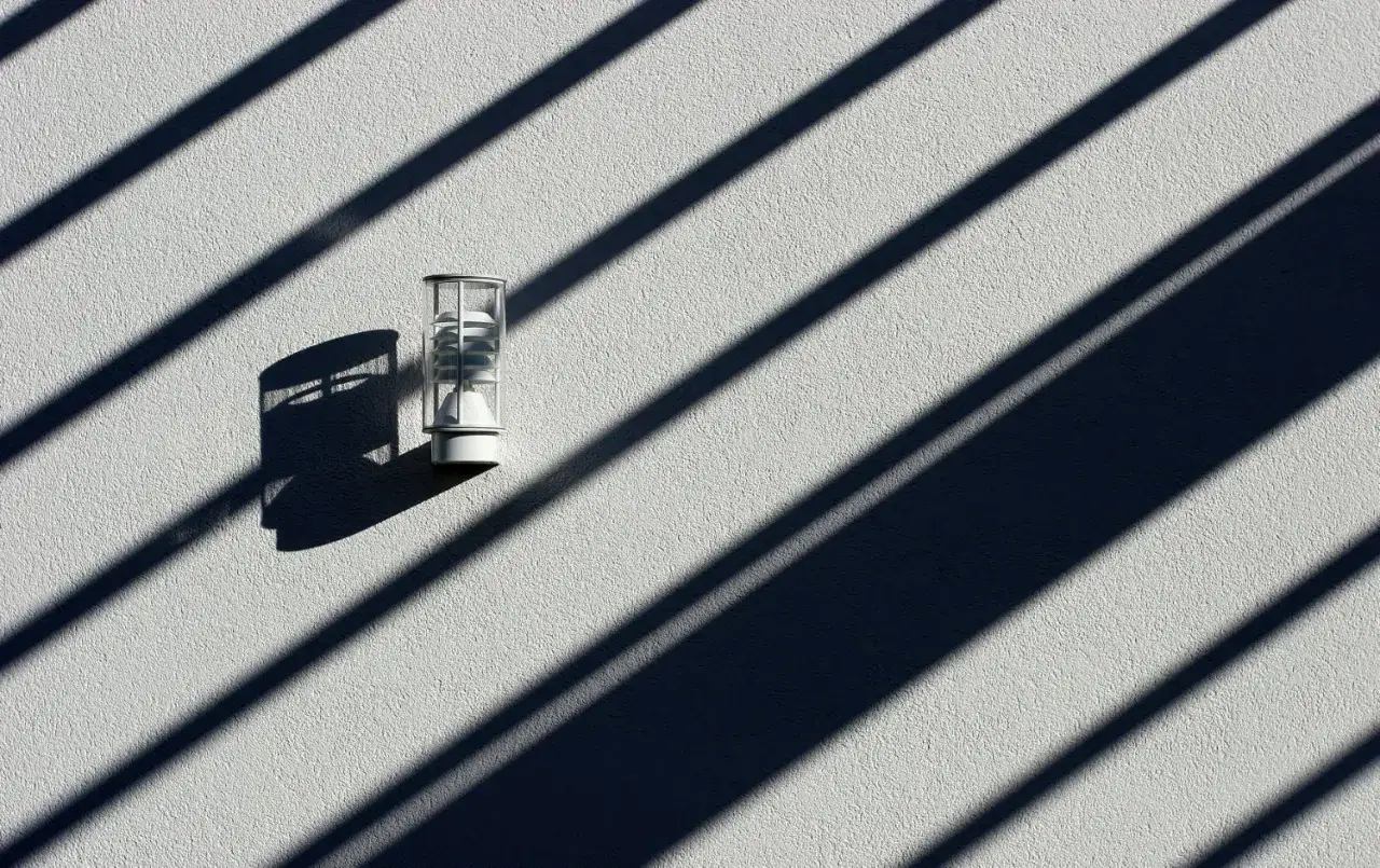 Een buitenlamp op een witte muur, met een dramatische diagonaal compositie van schaduwen van lamellen.