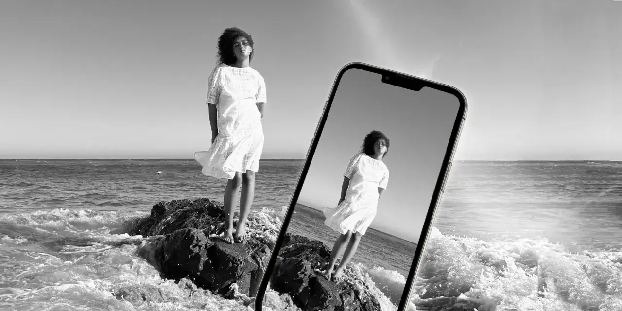 Zwart-wit foto van een vrouw in een witte jurk op rotsen bij de zee, weerspiegeld in een iPhone.