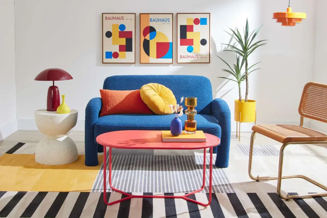 Een woonkamer met blauwe bank, rode salontafel en drie posters met bauhaus kunst. Gele en oranje kussens.