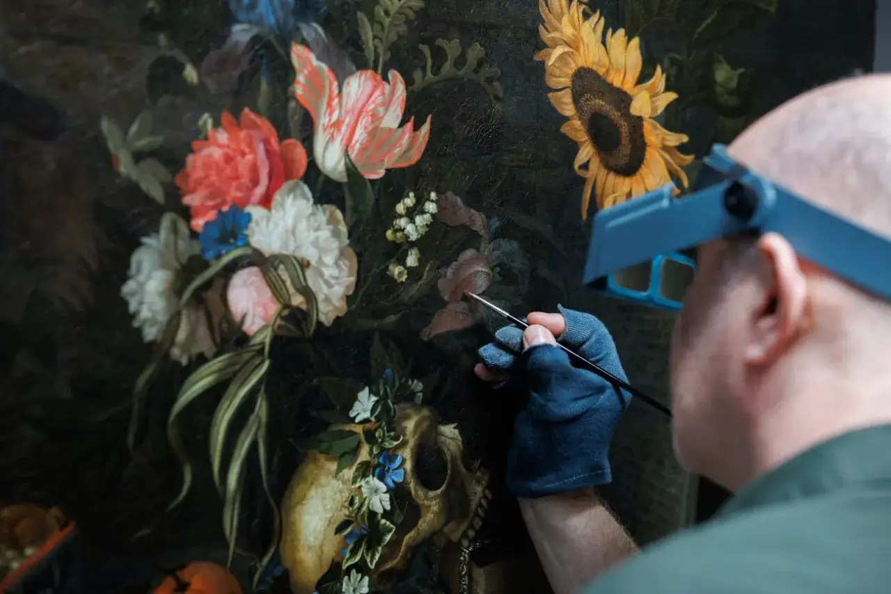 Restaurateur werkt aan een bloemstillevenschilderij, mogelijk van Maria van Oosterwijck. De details van de bloemen en schedel worden met precisie hersteld.