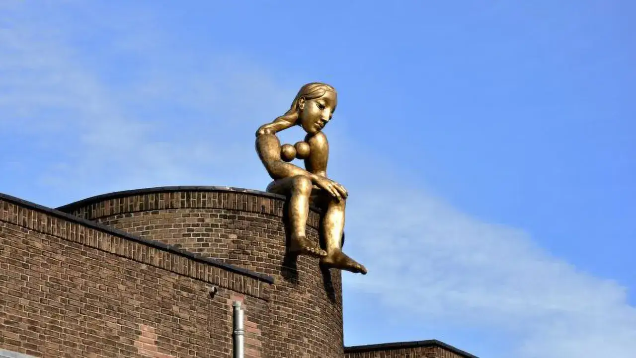 Gouden beeld van een zittende vrouw, die over de stad uitkijkt. Ze lijkt te mijmeren, een plassende vrouw in gedachten.