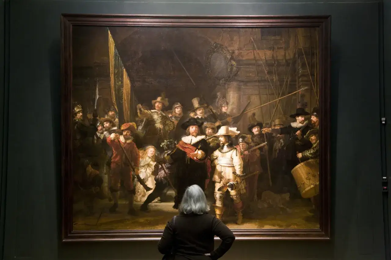 Een vrouw kijkt naar De Nachtwacht, een beroemd schilderij van Rembrandt. De dynamische compositie toont een groep schutters in actie.