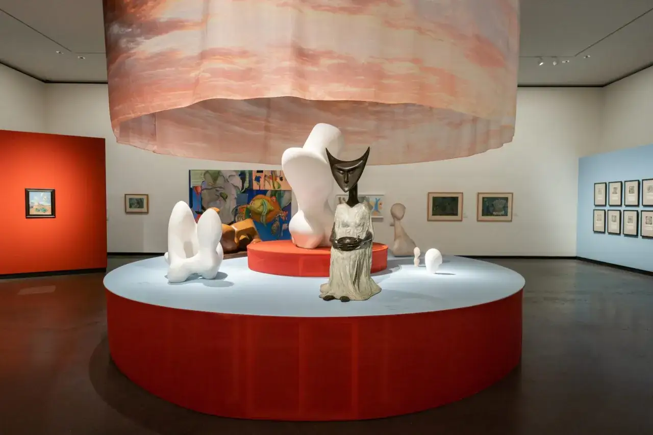 Een museumzaal met surrealisme kunstwerken, waaronder beelden en schilderijen, onder een zwevende, wolkenachtige stof.
