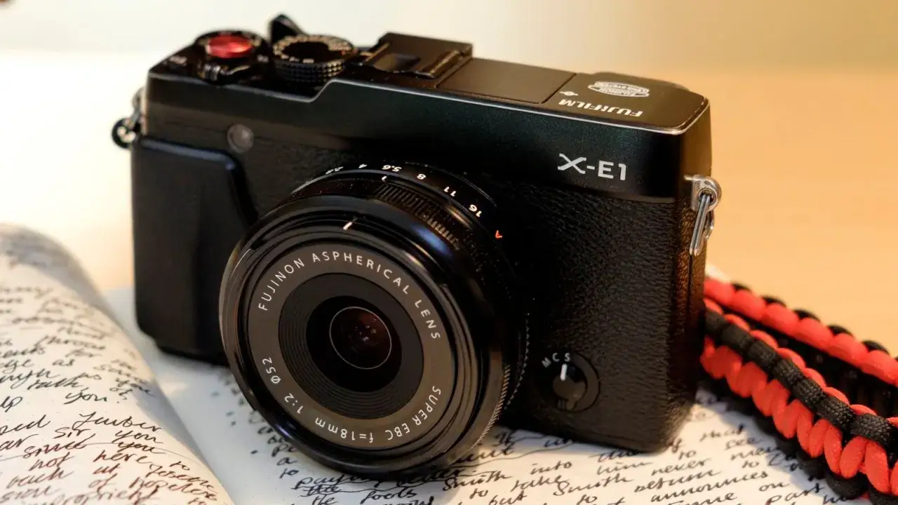 Zwarte Fujifilm X-E1 camera met lens, op een open schrift. De camera heeft een rode ontspanknop en een rode en zwarte draagband.