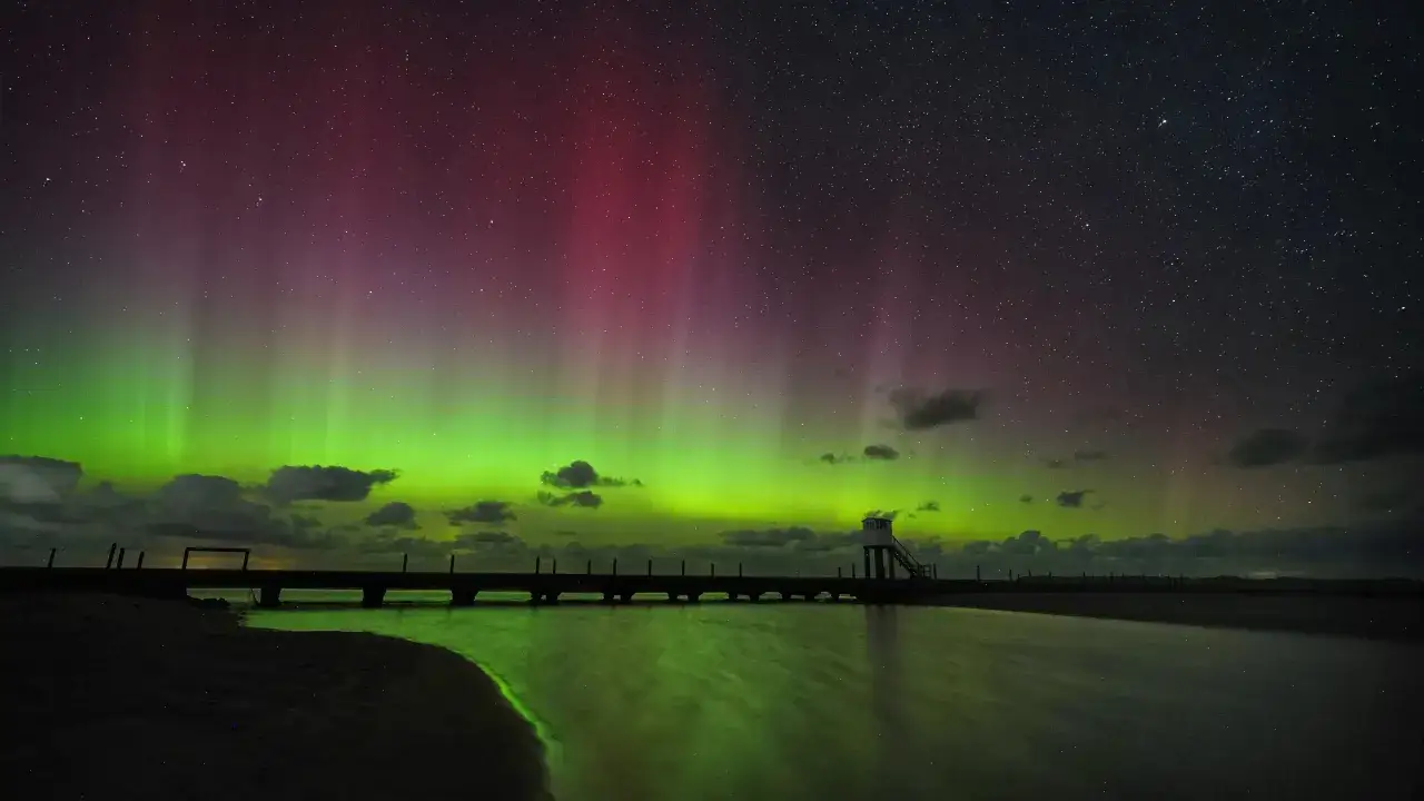 Spectaculaire foto's van het noorderlicht boven Nederland. Groene en rode gordijnen dansen aan de sterrenhemel boven een pier en strand.