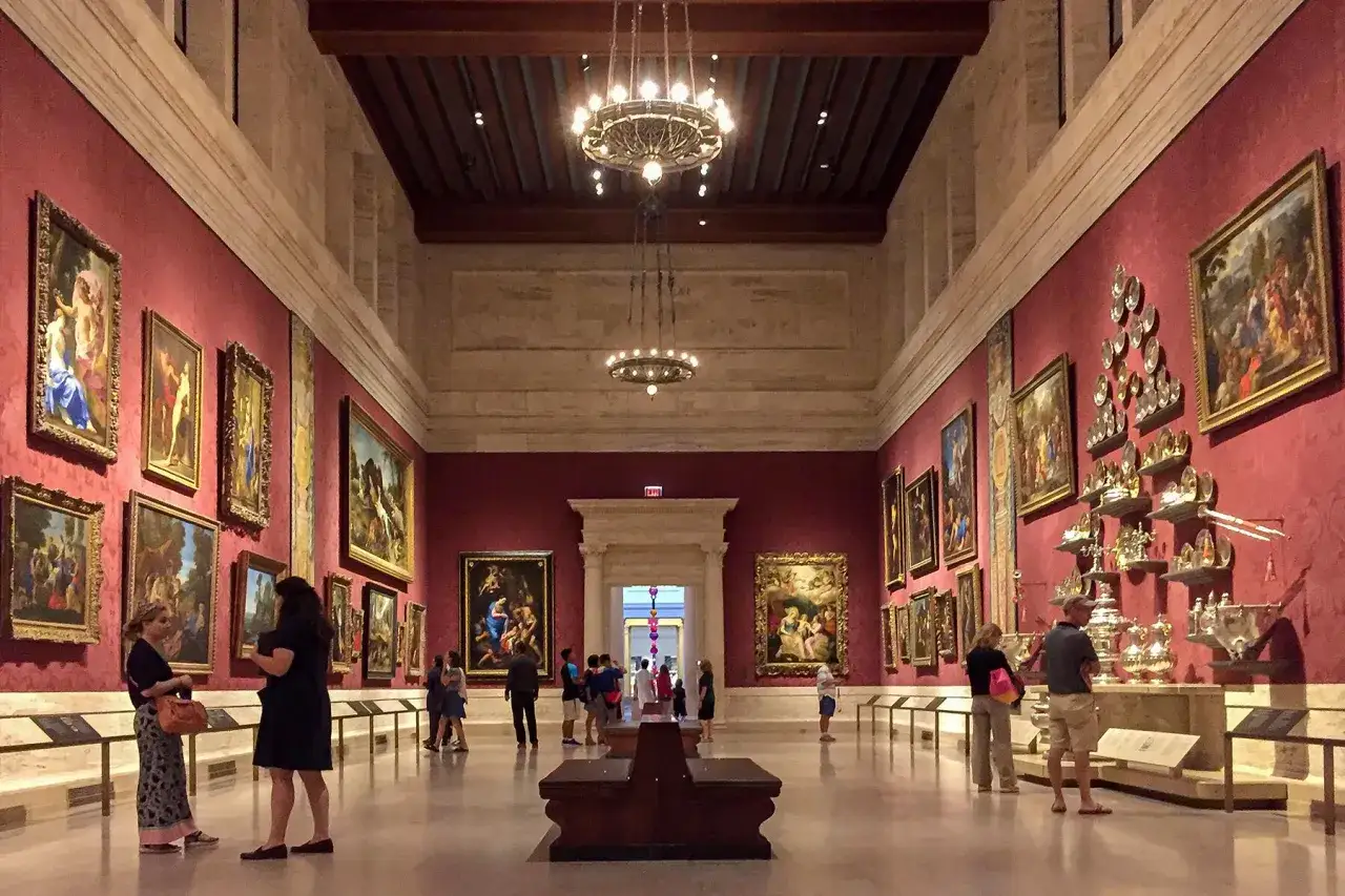 Een museumzaal met schilderijen van bekende kunstenaars aan de rode muren. Bezoekers bewonderen de kunstwerken, onder de indruk van de meesterwerken.