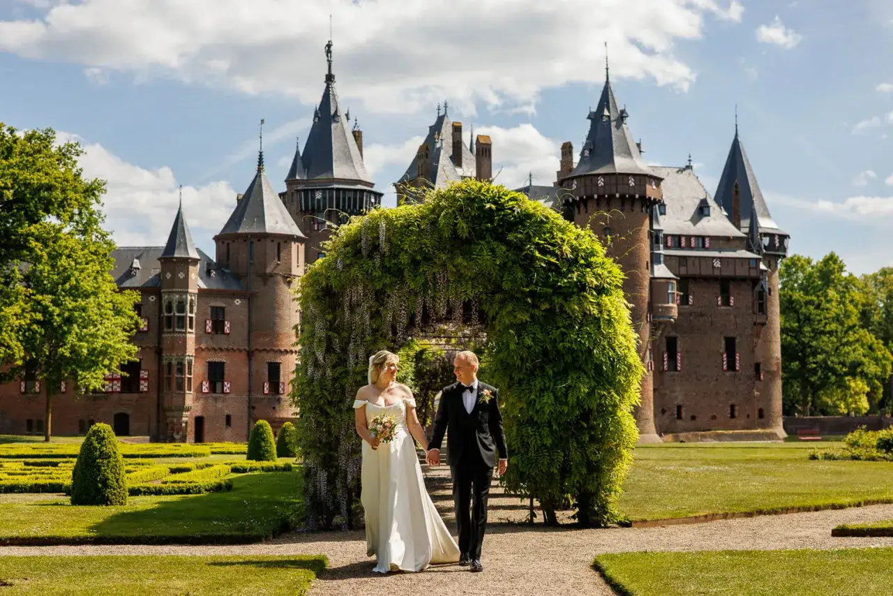 Bruidspaar loopt hand in hand onder een bloeiende boog door, met een sprookjesachtig kasteel op de achtergrond. Een perfecte fotoshoot kasteel.