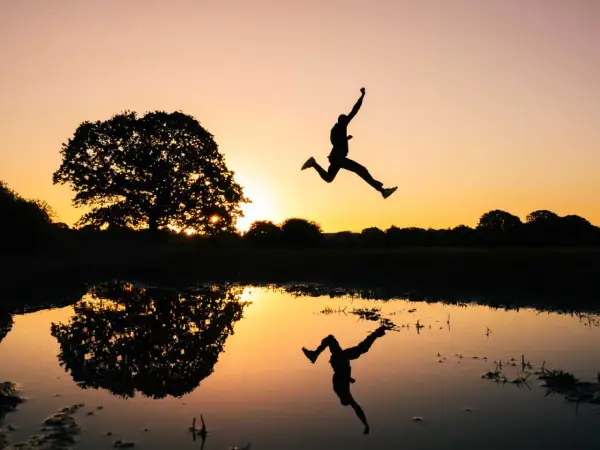 Silhouet van een persoon die springt bij zonsondergang, met een reflectie in het water. Een perfecte burst foto Samsung.
