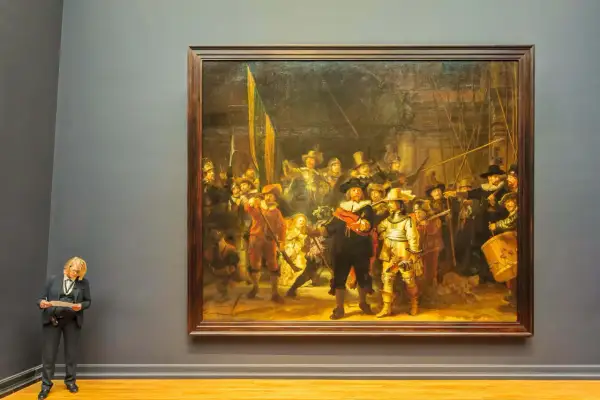 Een vrouw met blond haar en bril staat voor het beroemde nachtwacht schilderij, met een document in haar handen.