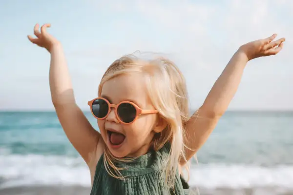 Vrolijk meisje met zonnebril op het strand, armen in de lucht. Ze lijkt wel een kleine godin, met namen als Freya of Aphrodite.