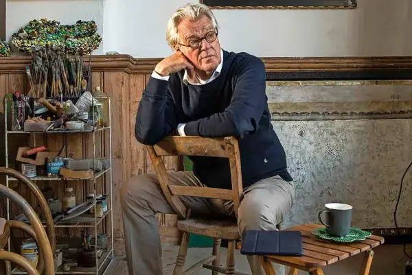 Jeroen Krabbé zit peinzend op een stoel in zijn atelier, omringd door schildersbenodigdheden.