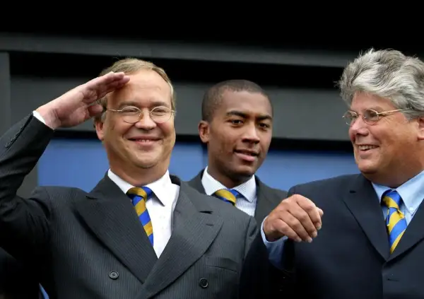 Pim de Lange salueert met een brede glimlach, naast twee mannen in pak.
