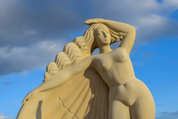 Zandstenen beeld van de Griekse godin Aphrodite, met golvend haar en een hand op haar hoofd, tegen een blauwe lucht met wolken.
