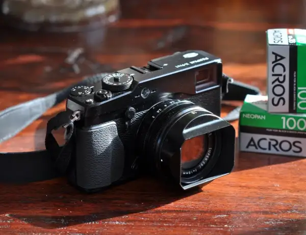 Zwarte Fujifilm X-E1 camera met lenskap en filmrolletjes Acros 100 op een houten tafel.